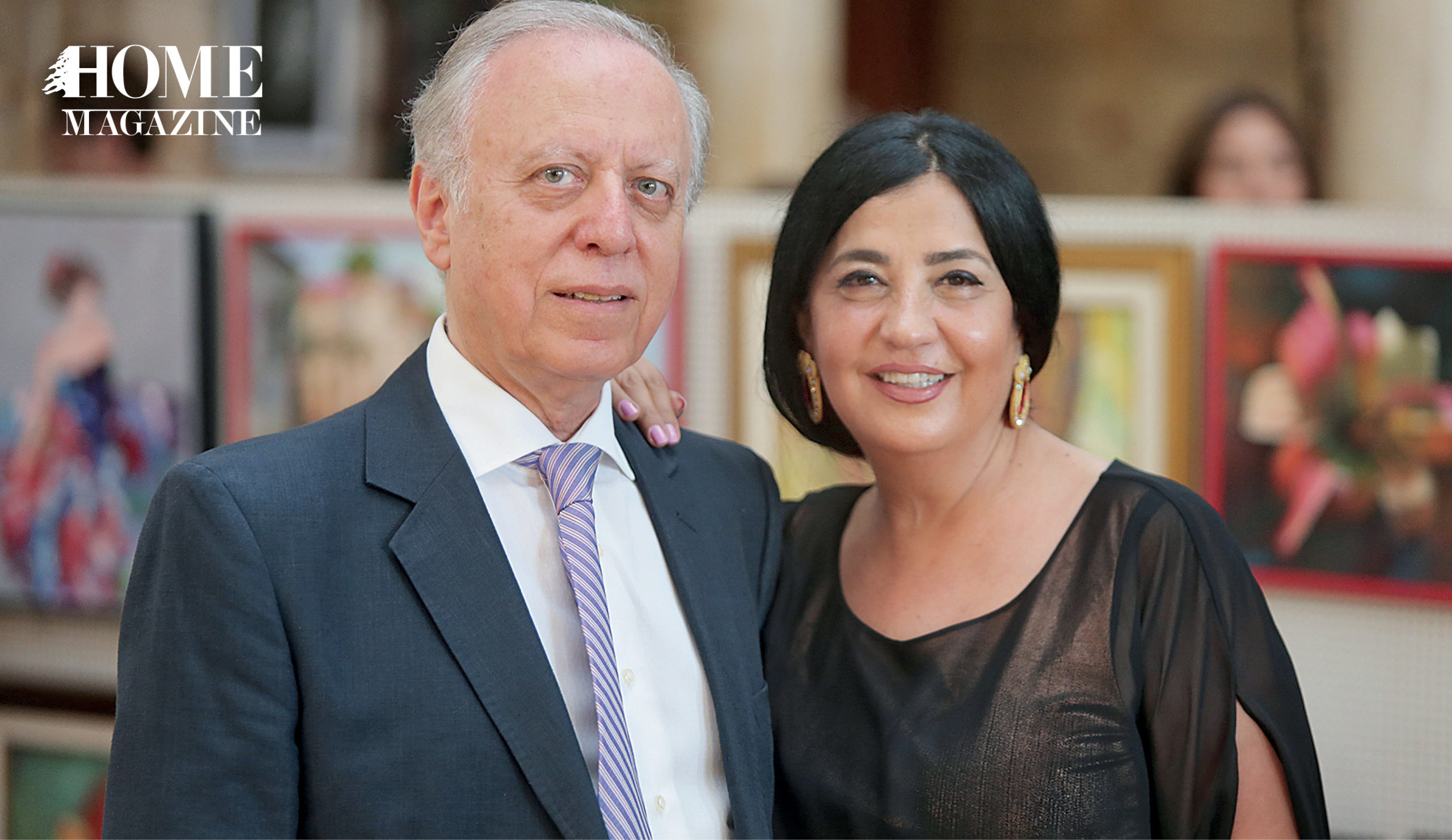 Man of white hair in suit and woman of black hair in black attire with paintings in the background