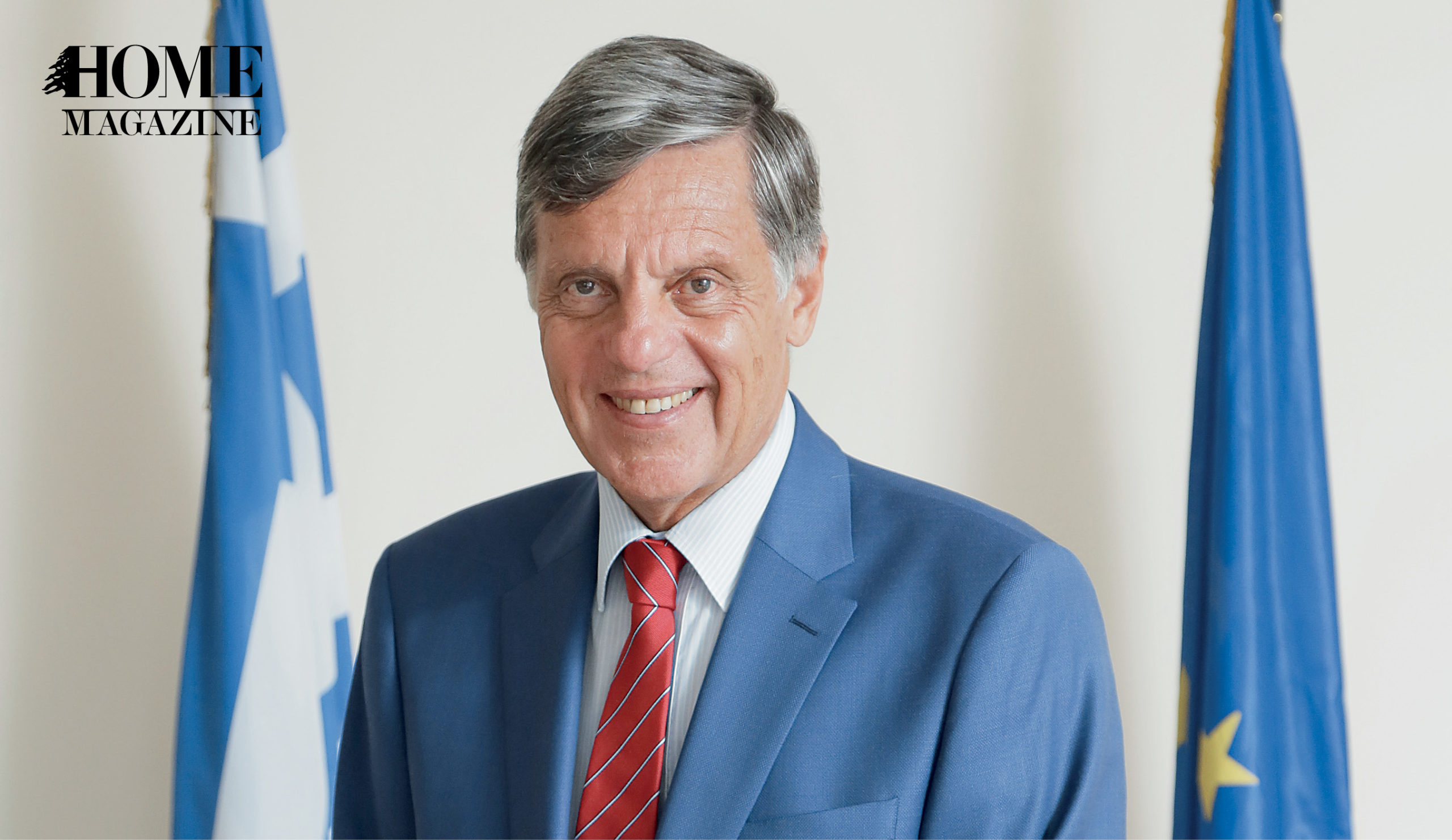 Smiling old man wearing navy suit and red tie with two blue flags in background